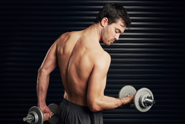 Construindo o corpo perfeito Foto de estúdio de um homem sem camisa e bem construído levantando halteres aquecendo contra um fundo escuro