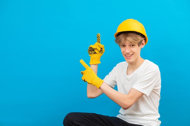 Construtor adolescente bonito feliz em um capacete amarelo está olhando para a câmera e aponta os dedos para cima em pé em um estúdio em um fundo azul