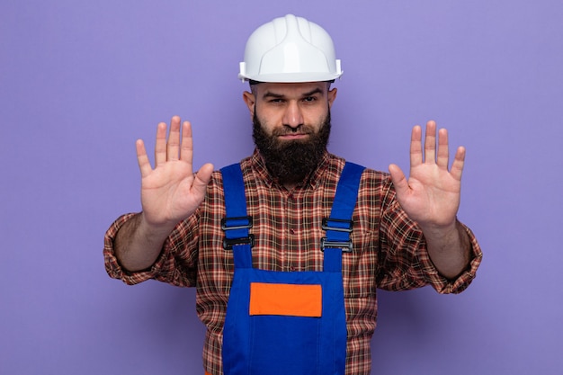 Construtor barbudo com uniforme de construção e capacete de segurança, olhando com uma cara séria, fazendo gesto de parada com as mãos