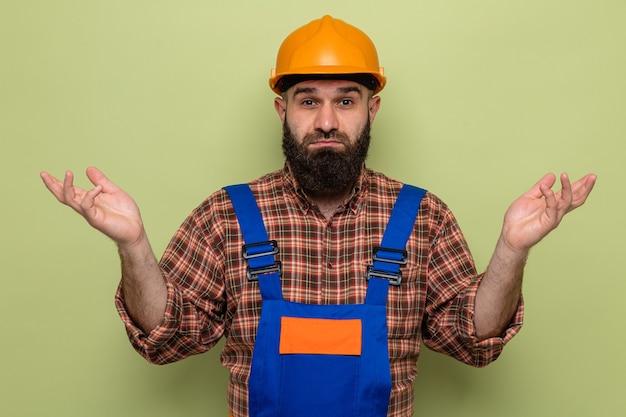 Construtor barbudo com uniforme de construção e capacete de segurança, olhando para a câmera, confuso, encolhendo os ombros e sem resposta em pé sobre um fundo verde