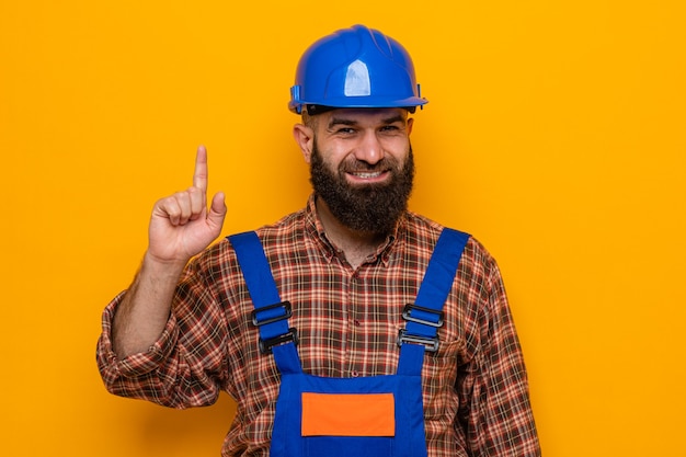 Construtor barbudo em uniforme de construção e capacete de segurança olhando para a câmera feliz e alegre, mostrando o dedo indicador tendo uma ótima ideia em pé sobre um fundo laranja