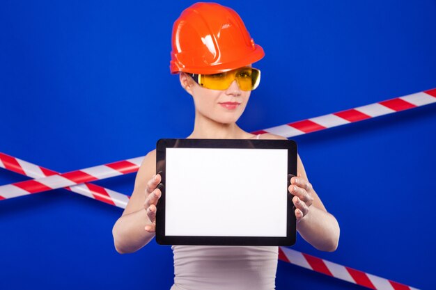 Construtor bonita mulher de camisa branca, cinto de construtor, capacete, óculos de construtor, shorts jeans e snickers segurar tablet vazio