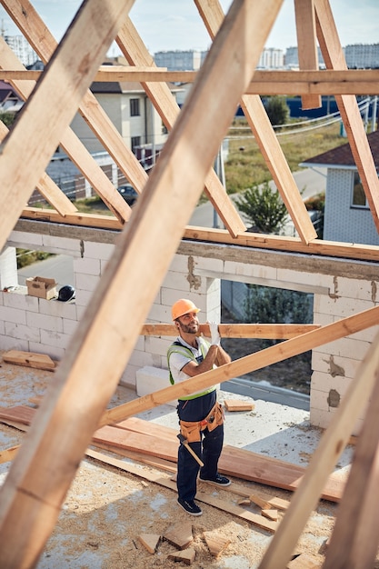 Construtor de aparência positiva segurando um longo pedaço de madeira de construção enquanto faz uma carcaça de telhado