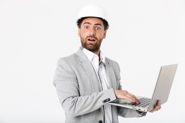 Construtor de homem barbudo confuso, vestindo terno e capacete de segurança, isolado na parede branca, mostrando o computador laptop