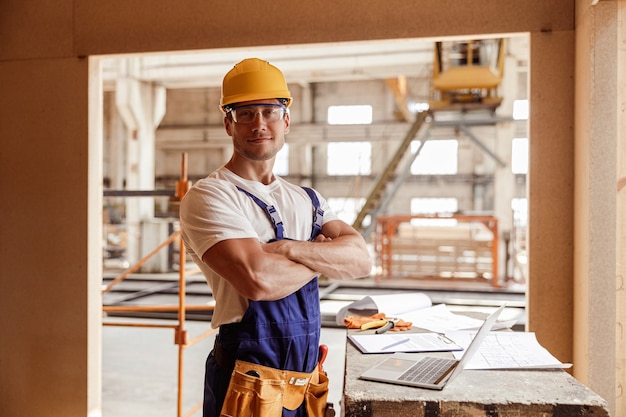 Construtor de homem bonito em pé ao lado da mesa com um laptop