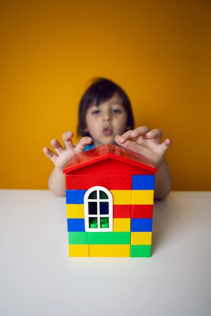 Construtor de menino criança em uma camiseta azul contra uma parede amarela está construindo uma casa de cubos de plástico coloridos.