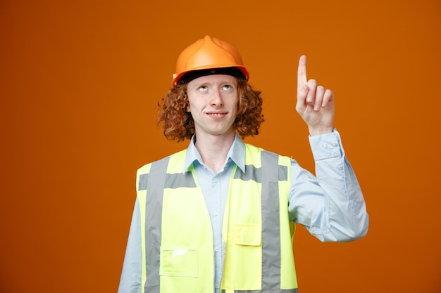 Construtor jovem em uniforme de construção e capacete de segurança olhando para cima sorrindo apontando com o dedo indicador para cima em pé sobre fundo laranja