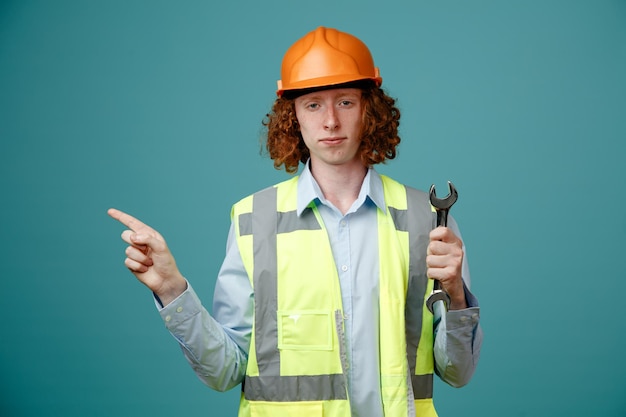 Construtor jovem em uniforme de construção e capacete de segurança segurando a chave apontando com o dedo indicador para o lado olhando confiante em pé sobre fundo azul