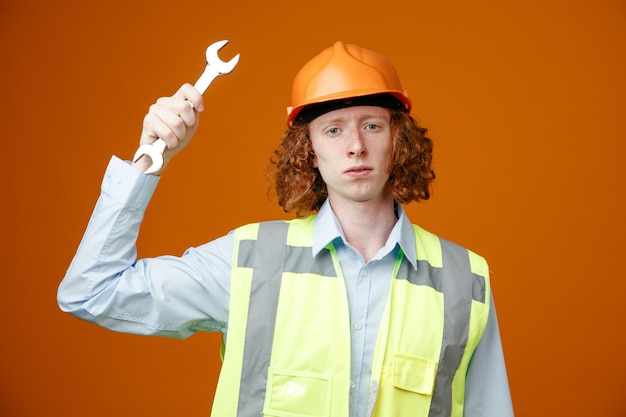 Construtor jovem em uniforme de construção e capacete de segurança segurando a chave olhando para a câmera com cara séria em pé sobre fundo laranja