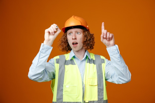 Construtor jovem em uniforme de construção e capacete de segurança segurando o marcador parecendo surpreso apontando com o dedo indicador para cima tendo ótima idéia em pé sobre fundo laranja