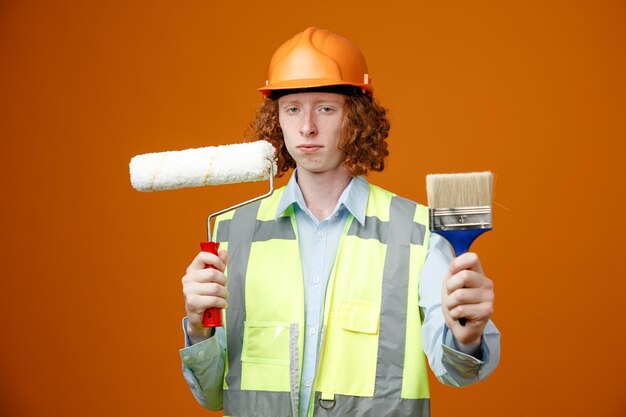 Construtor jovem em uniforme de construção e capacete de segurança segurando o rolo de pintura e pincel olhando para a câmera com cara séria em pé sobre fundo laranja