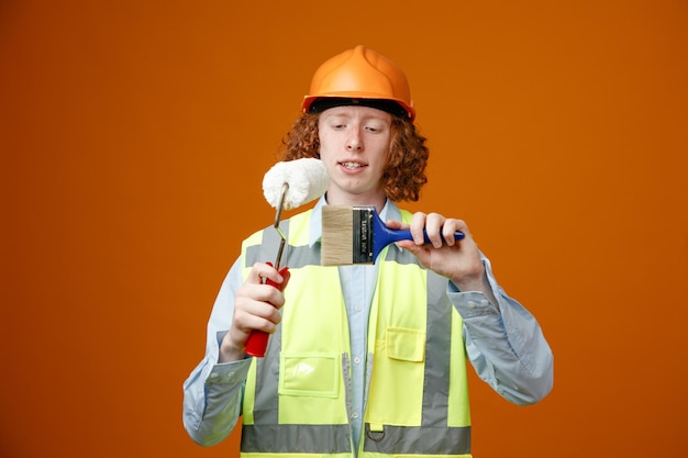 Construtor jovem em uniforme de construção e capacete de segurança segurando rolo de pintura e pincel olhando intrigado sorrindo em pé sobre fundo laranja