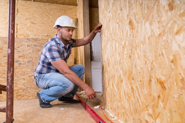 Construtor masculino, trabalhador da construção civil, usando capacete branco, medindo a moldura da porta com fita métrica dentro de casa inacabada com placas de madeira compensada de partículas expostas