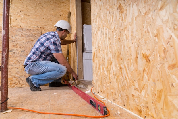 Construtor masculino, trabalhador da construção civil, usando capacete branco, medindo a moldura da porta com fita métrica dentro de casa inacabada com placas de madeira compensada de partículas expostas