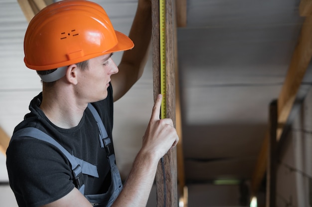 Construtor masculino vestido com roupa de trabalho e um capacete laranja. Mede a distância com uma fita métrica