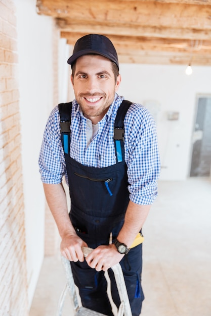 Foto construtor sorridente em pé na escada no espaço de trabalho dentro de casa