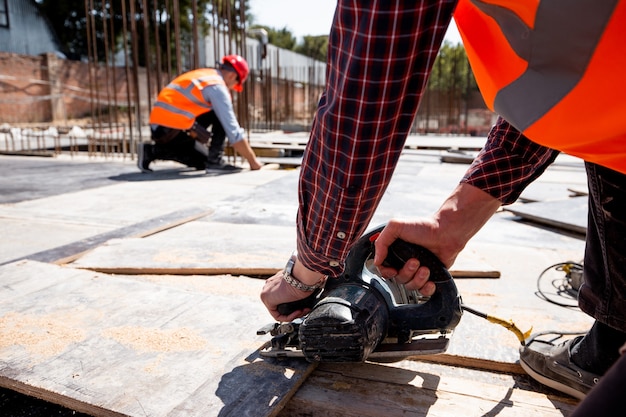 Construtor vestido com colete de trabalho laranja e capacete está usando uma serra circular no canteiro de obras aberto. .