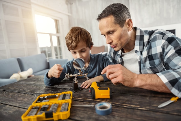 Construyendo. Apuesto hombre de cabello oscuro amoroso concentrado mostrando instrumentos a su hijo mientras está sentado en la mesa y su hijo mirando una lupa