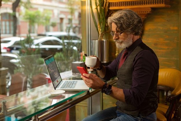 Construyendo contactos. Grave hombre adulto sentado en la mesa de café con su computadora portátil escribiendo un mensaje y tomando café durante su jornada laboral.