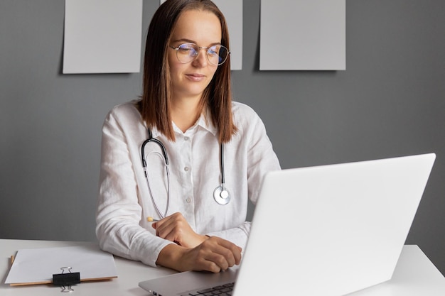 Consulta on-line seguro de pesquisa bela jovem médica vestindo bata de laboratório sentada na frente de computador portátil digitando no teclado tendo chamada de vídeo com paciente on-line