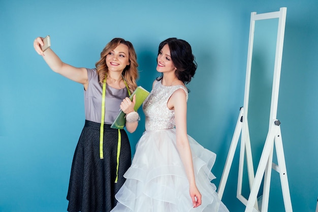 Consultor de boda costurera y novia probándose un vestido de novia y hacer selfie en el estudio sobre un fondo azul. sastre, modista, y, cliente, prom, en, el, vestidor