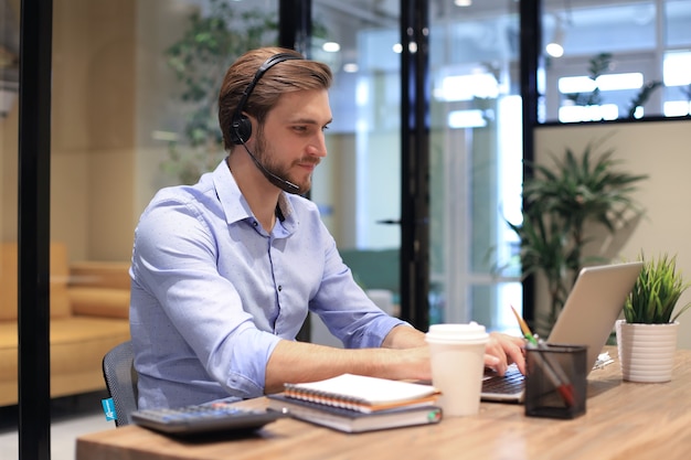 Consultor de negócios masculino sorridente com fones de ouvido sentado no escritório moderno, videochamada, olhando para a tela do laptop. Linha de apoio do agente de suporte de atendimento ao cliente do homem falando no chat online.