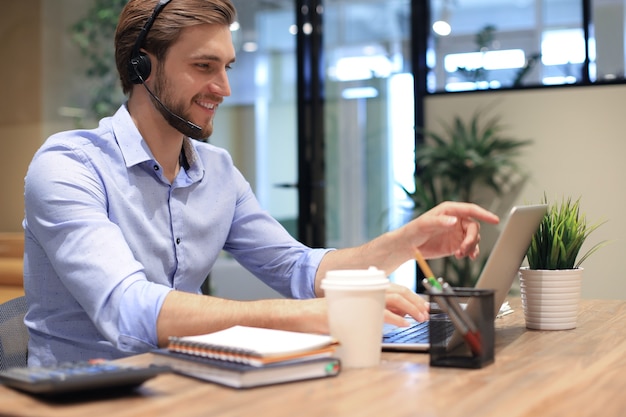 Consultor de negócios masculino sorridente com fones de ouvido sentado no escritório moderno, videochamada, olhando para a tela do laptop. Linha de apoio do agente de suporte de atendimento ao cliente do homem falando no chat online.