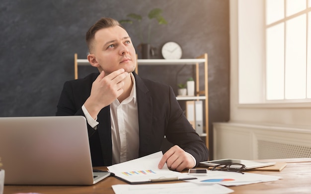 Consultor financeiro masculino sério, trabalhando com papéis e laptop no interior do escritório moderno. Homem pensativo com roupa formal, verificando documentos e pensando sobre a estratégia, copie o espaço