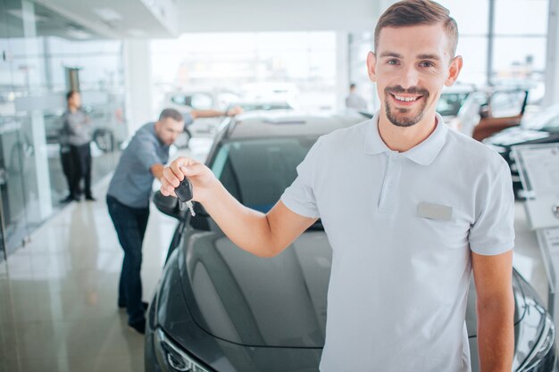 Consultor jovem e barbudo fica e detém a chave do carro preto. Ele olha para caera e sorri. Cara veste camisa branca. O potencial comprador está próximo. Ele olha para dentro do carro.