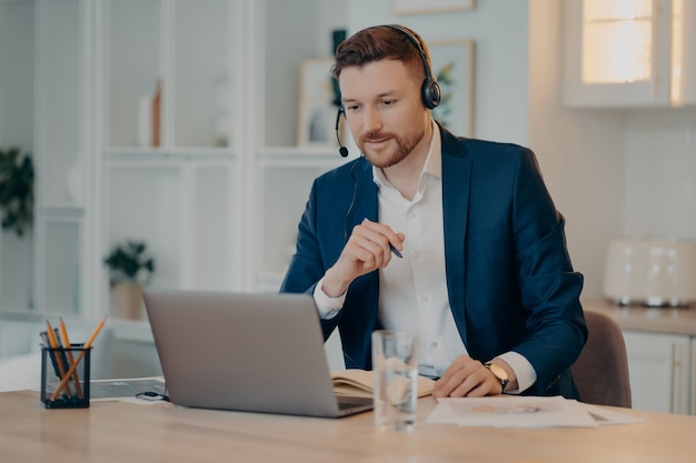 Consultor de negocios masculino alegre en auriculares trabajando en casa mientras habla con su socio o cliente durante una reunión en línea o una videollamada en la computadora portátil, sentado en su lugar de trabajo. Negocios en línea