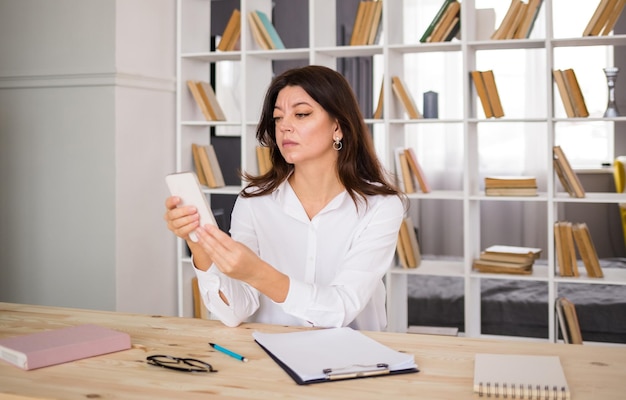 Una consultora adulta con una blusa blanca está sentada en una mesa en una habitación y hablando por teléfono Trabajar en casa