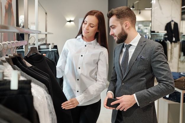Consultora de moda morena en blusa blanca mostrando suéter al hombre mientras le da consejos en la tienda de ropa