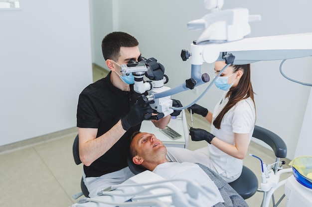 Consultorio dental moderno con un microscopio Un dentista trata los dientes de un hombre bajo un microscopio un asistente lo ayuda Atención médica moderna para el dolor de muelas