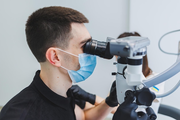 Consultório odontológico moderno com um microscópio Um dentista trata os dentes de um homem sob um microscópio um assistente o ajuda Cuidados médicos modernos para dor de dente