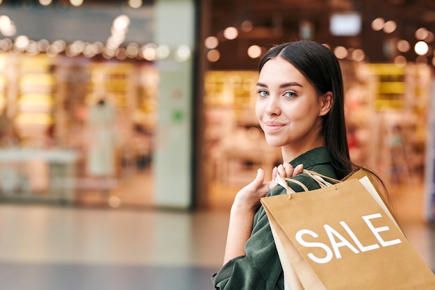 Consumidor bastante joven con un montón de bolsas de papel de pie delante de la cámara mientras visita un gran centro comercial moderno durante la temporada de rebajas