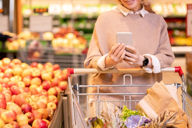 Consumidor de hembras maduras en jersey beige tejido empujando el carro con productos alimenticios en supermarker y desplazamiento en smartphone