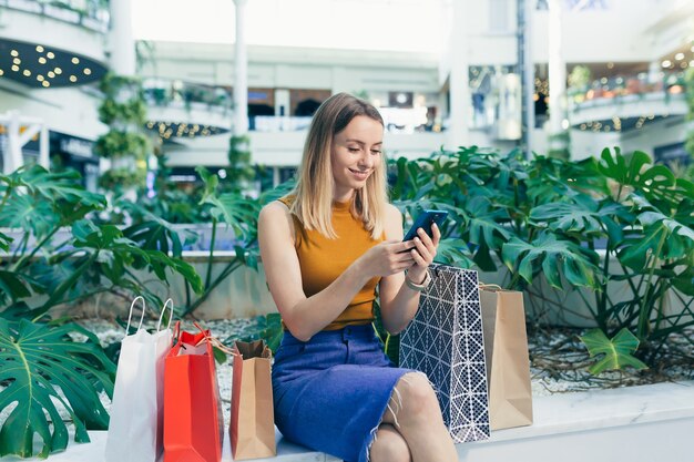 Consumidor jovem no shopping navega no bate-papo e usa um smartphone. mulher em pé com um telefone celular nas mãos em shopping center. interior. compradora feliz com sacolinhas fazendo compras