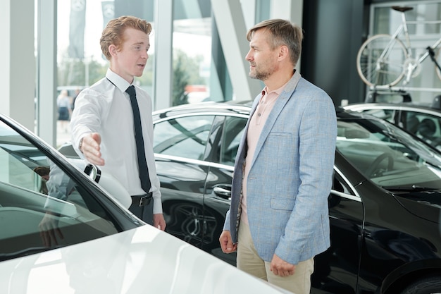 Consumidor masculino maduro en ropa casual mirando al joven gerente de ventas apuntando al coche blanco y escuchándolo mientras elige un nuevo auto