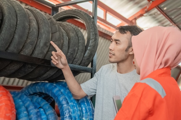 Consumidor masculino olha para um pneu apontando um dedo selecionando um pneu com uma mecânica velada em uma oficina de peças sobressalentes de motocicletas