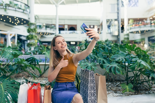 La consumidora joven en el centro comercial navega por el chat y utiliza un teléfono inteligente. mujer de pie con un teléfono móvil en sus manos en el centro comercial. interior. Compradora feliz niña con bolsas de regalo hacer compras