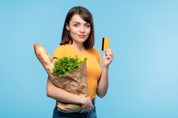 Consumidora muito jovem com uma sacola de papel contendo pão fresco e salsa verde mostrando seu cartão de crédito