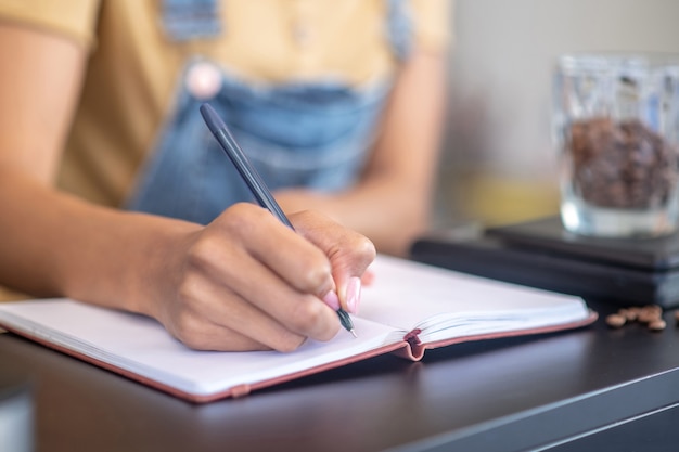 Contabilidade. Mão fina graciosa de mulher escrevendo com caneta no caderno de peso de grãos de café no balcão