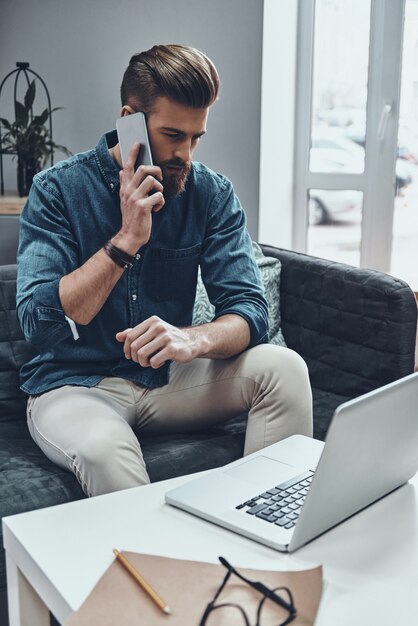 En contacto con clientes. Hombre moderno joven pensativo hablando por teléfono inteligente mientras está sentado en su lugar de trabajo