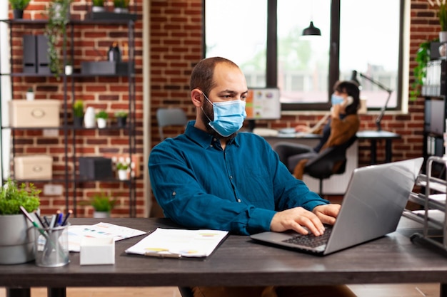 Foto contador con mascarilla médica de protección contra covid19 sentado en el escritorio escribiendo estrategia de marketing trabajando en la inversión de la empresa en la oficina de inicio. empresario analizando gráficos de negocios