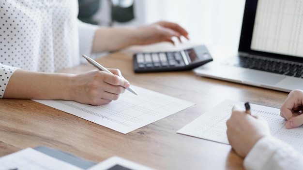 Contadora usando uma calculadora e um computador portátil enquanto conta impostos com um colega em uma mesa de madeira no escritório. Trabalho em equipe em auditoria e finanças de negócios.