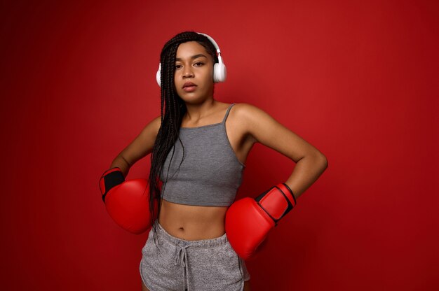 Contate o conceito da arte marcial. Jovem boxeador de mulher desportiva em fones de ouvido sem fio e roupas esportivas, coloca as mãos com luvas de boxe na cintura e olha para a câmera posando sobre fundo vermelho com espaço de cópia.