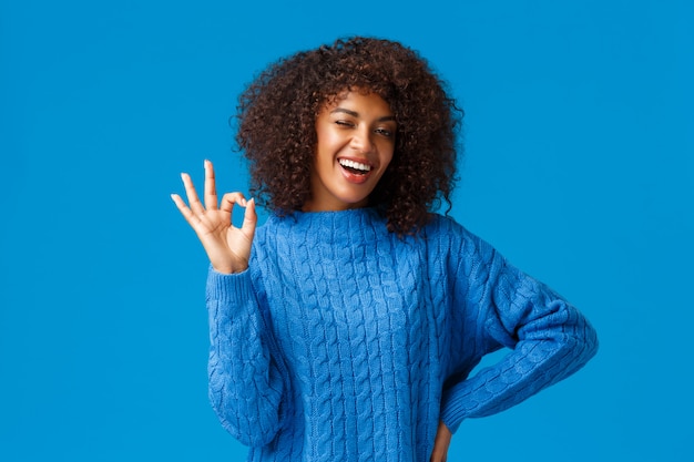 Conte comigo, está tudo bem. Alegre e assertiva garota afro-americana com corte de cabelo afro tem tudo sob controle, mostrando tudo bem gesto e piscadela, sorrindo resposta positiva, fundo azul