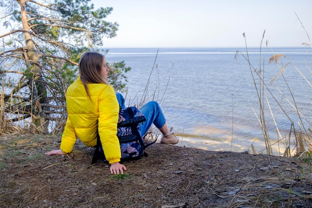 Contemplación mujer joven con mochila está sentada en la costa del lago del bosque en un día nublado en spri