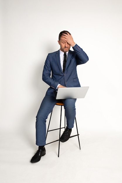 Contemplando o empresário se sentindo triste, o homem que trabalha no laptop. Gerente cansado sentado no estúdio em fundo branco com o computador portátil com aparência cansada, dor de cabeça, chocado, exausto. Cansaço.