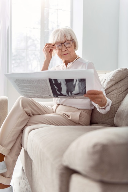 Contenido apasionante. Agradable anciana sentada en el sofá y leyendo un periódico interesante mientras ajusta sus anteojos